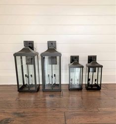 three black lanterns sitting next to each other on a wooden floor in front of a white wall