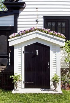 a black and white dog house with purple flowers on the roof, in front of a home