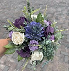 a bouquet of purple and white flowers is held by someone's hand on a brick walkway