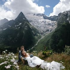 a man and woman sitting on top of a lush green hillside next to mountains covered in snow