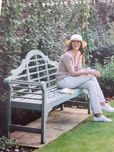 a woman sitting on a bench in front of a wall with plants and bushes behind her