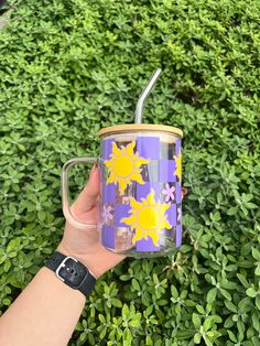 a hand holding a purple and yellow coffee mug in front of some green bushes with sunflowers on it