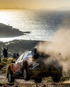 a car is driving down a dirt road near the ocean with people standing around it