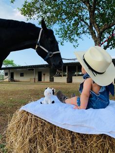 a baby in overalls and cowboy hat sitting on hay next to a black horse