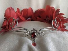 A beautiful, intricate crown with a ruby red crystal decorating the center of the silver wire and peonies in various shades of red surrounding the piece. Eyes will follow the delicate swirls and romantic peonies as the garland decorates your head, perfect for renaissance fairs, weddings, cosplay, or any event of your choosing! Red Crown Jewelry For Wedding, Silver Adjustable Crown With Pinched Shape, Red Crown Headpiece For Wedding, Elegant Handmade Wedding Crown, Elegant Teardrop Crown Headpiece For Gifts, Elegant Headpiece With Teardrop Crown For Gift, Elegant Headpieces With Teardrop Crown For Gift, Elegant Round Crown Headpiece As Gift, Elegant Round Crown Headpiece For Gifts