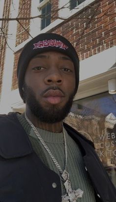 a man with a beard wearing a black jacket and a beanie is standing in front of a brick building