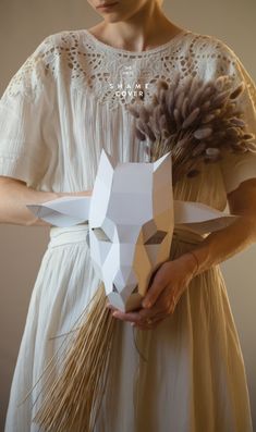 a woman in a white dress holding a paper animal mask with dried grass on it