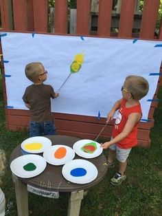 two young boys standing in front of a table with paper plates and paintbrushes on it