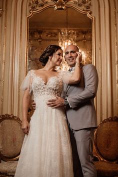 a bride and groom pose for a photo in front of an ornate gold framed mirror