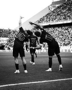 three men are playing soccer in front of an audience at a sporting event, one is reaching up for the ball