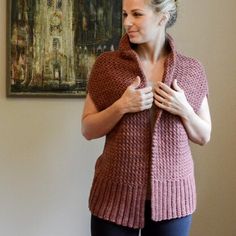 a woman standing in front of a painting wearing a pink knitted sweater and black pants