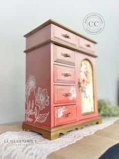 an old dresser painted pink and gold with flowers on the drawers is sitting on a lace tablecloth