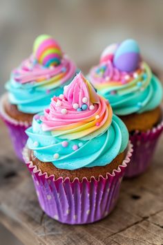 three cupcakes with blue frosting and rainbow icing on wooden table top