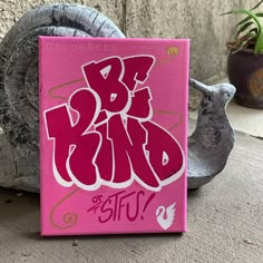 a pink canvas with the word be kind on it next to a bird statue and potted plant