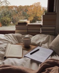 an open book, coffee cup and pen on top of a bed in front of a window