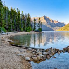 a lake surrounded by trees and rocks with mountains in the background on a sunny day