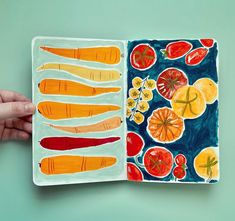 a hand holding an open book with fruit and vegetables painted on it