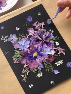 a hand holding a blue and purple flower in front of a black card with flowers on it