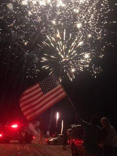 fireworks and an american flag are lit up in the night sky with people standing on the side of the road