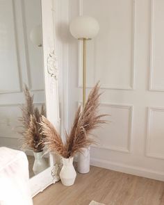 a white vase filled with dry grass sitting on top of a wooden floor next to a mirror