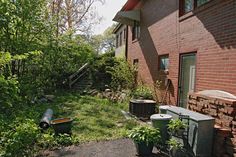 an overgrown back yard with trash cans and garbage cans in the foreground, next to a brick building