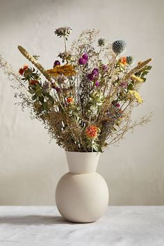 a white vase filled with lots of wild flowers on top of a table next to a wall