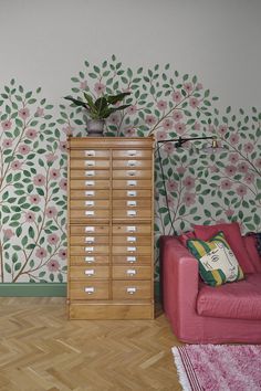 a living room with floral wallpaper and pink chair next to a wooden filing cabinet