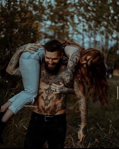 a tattooed man carrying a woman on his back in a field with trees behind him