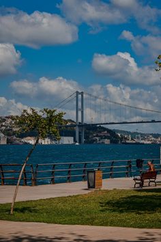 people are sitting on benches near the water and under a bridge with a view of the city