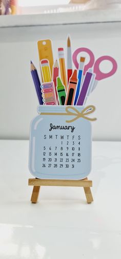 a desk calendar with markers, scissors and pencils in it on a wooden stand