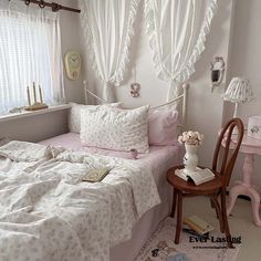 a white bed sitting next to a window covered in curtains and pink comforter on top of a wooden chair