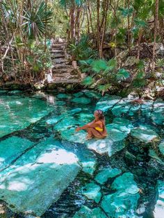 a woman sitting on top of a rock in the middle of a river surrounded by trees