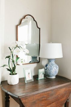 a wooden table topped with a mirror and vase filled with flowers next to a lamp