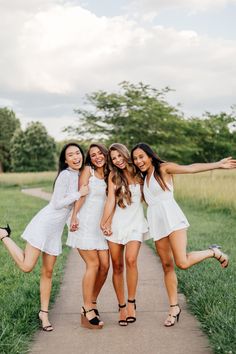 four girls in white dresses are posing on a path with their arms around each other