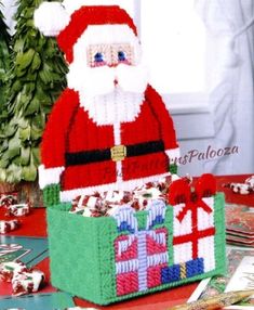 a knitted santa clause sitting in front of a box filled with presents on top of a table