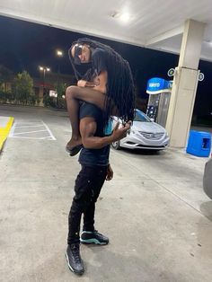 a man holding a woman in his arms at a gas station with cars parked behind him