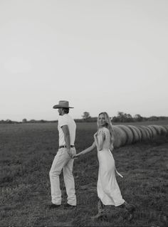 a man and woman holding hands while standing in the middle of a field with hay bales behind them