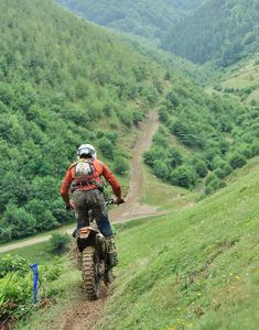 a man riding on the back of a motorcycle down a dirt road in front of a lush green hillside