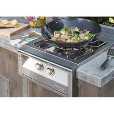 a large frying pan sitting on top of an outdoor gas stove next to a cutting board and utensils