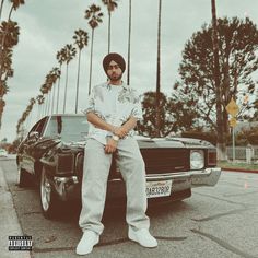 a man sitting on the hood of a car in front of palm trees with his arms crossed