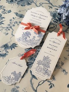 two tags with red ribbon tied to them sitting on a floral print table cloth next to blue and white flowers