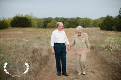 an older couple holding hands walking down a dirt path in the middle of a field