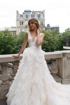 a woman in a white wedding dress standing on a stone balcony with trees and buildings behind her