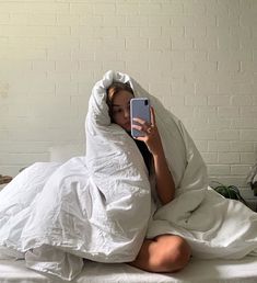 a woman sitting on a bed under a blanket holding a cell phone up to her face