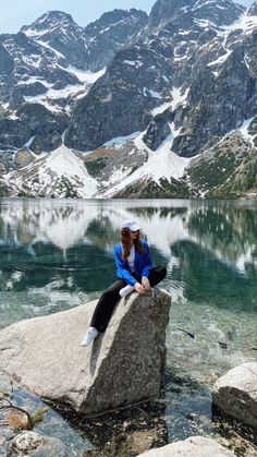 a woman sitting on top of a rock next to a body of water with snow covered mountains in the background