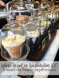 several cupcakes in glass cases on a counter top with other desserts behind them