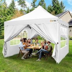 a group of people sitting around a table under a white tent on top of a grass covered field