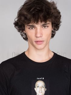 a young man with curly hair wearing a black t - shirt and looking at the camera