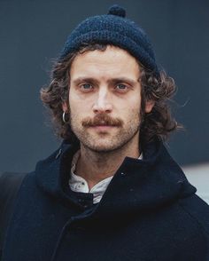 a man with curly hair wearing a blue hat and coat looks at the camera while standing in front of a building