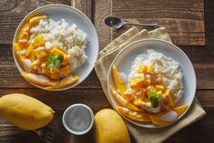 two white plates topped with rice and mangoes next to some fruit on top of a wooden table